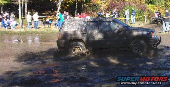 Mud-covered Jeep Grand Cherokee driving through a muddy track with spectators in the background