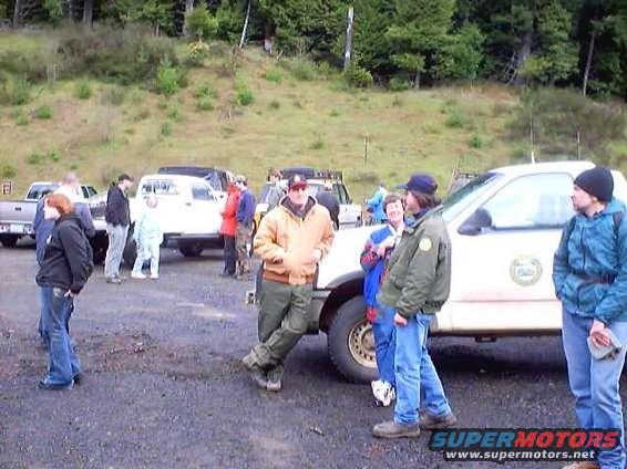 Group of people gathered near several trucks and SUVs in a wooded area