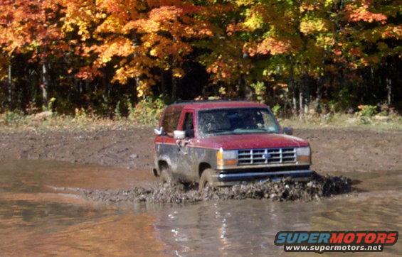 Red and black SUV with a blue grille driving through a muddy puddle, autumn trees in background