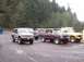 1976 Ford F-100 - Several Ford trucks and SUVs are parked in a gravel lot with trees in the background.