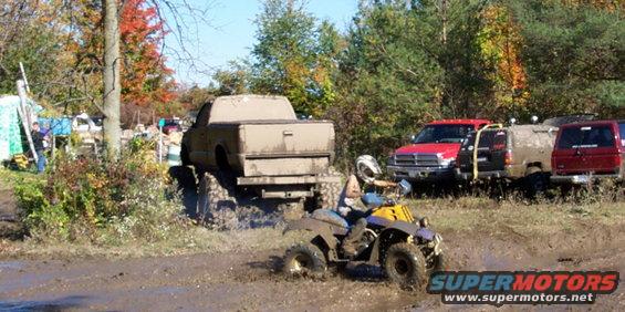 ATV splashing mud in front of a lifted pickup truck and other off-road vehicles in a muddy field