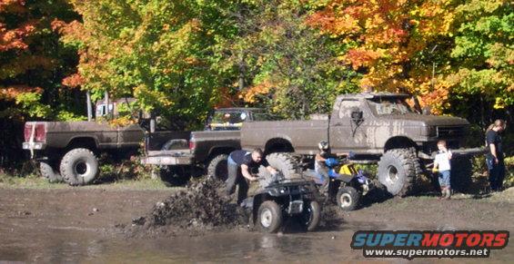 Several muddy pickup trucks and ATVs are in a wooded area with fall foliage. People are interacting with the vehicles in the mud