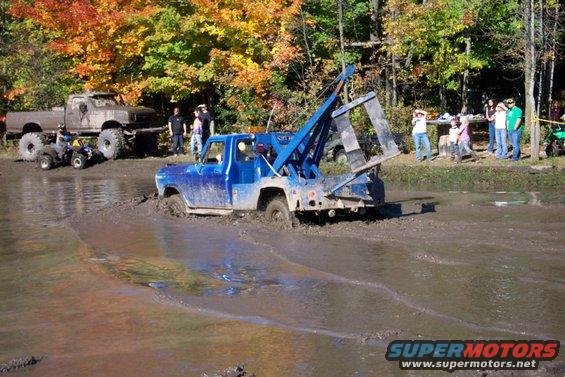 Blue tow truck stuck in mud pit with people watching. Another lifted truck and ATV are nearby