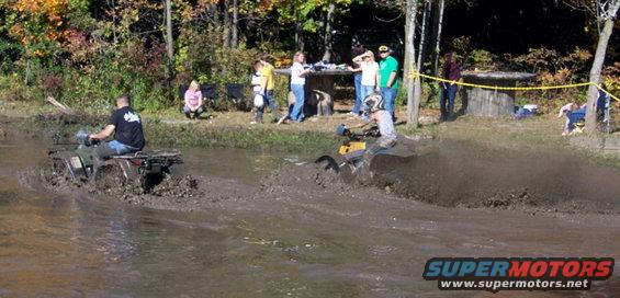 Two ATVs splashing through a muddy track with spectators watching from the bank