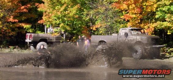 Two lifted trucks splashing mud while driving through water; autumn trees in background