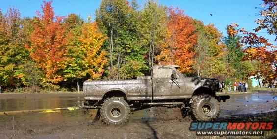 Muddy pickup truck with big tires driving through a mud pit with fall foliage in the background