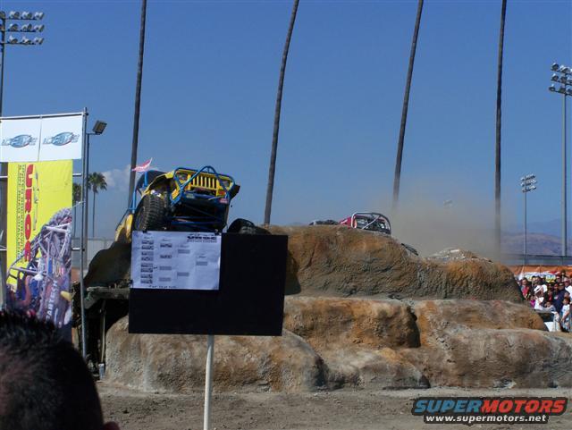 Yellow and blue off-road buggy on rocky terrain, with a second vehicle in the background kicking up dust