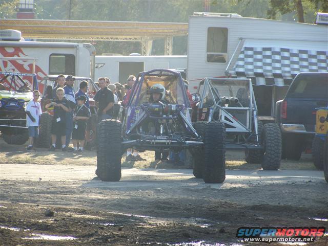 Buggy with roll cage and big tires, people around, trailers in background