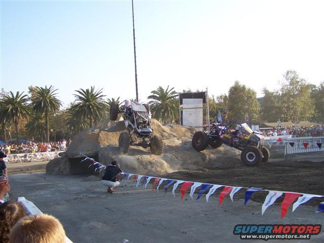 Off-road buggy with purple and yellow graphics climbing a dirt mound, with another buggy in the background