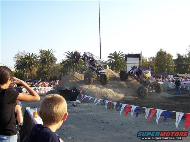 Two off-road buggies climbing dirt mounds at an expo
