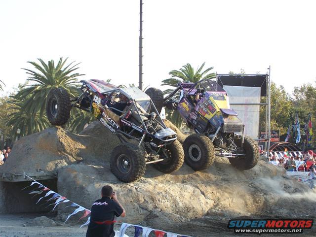 Two custom off-road vehicles climbing a dirt obstacle course with spectators in the background