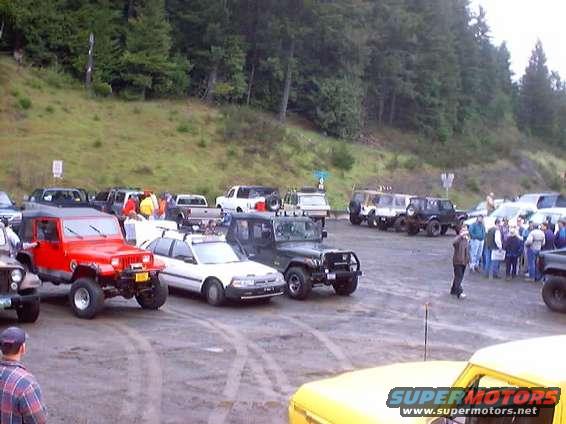 Group of off-road vehicles parked on dirt, with trees in background. People are gathered near some vehicles