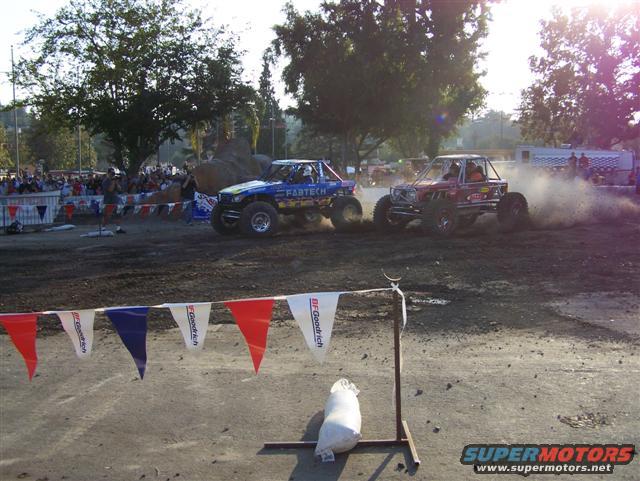 Two off-road buggies kicking up dust while racing, with spectators in the background