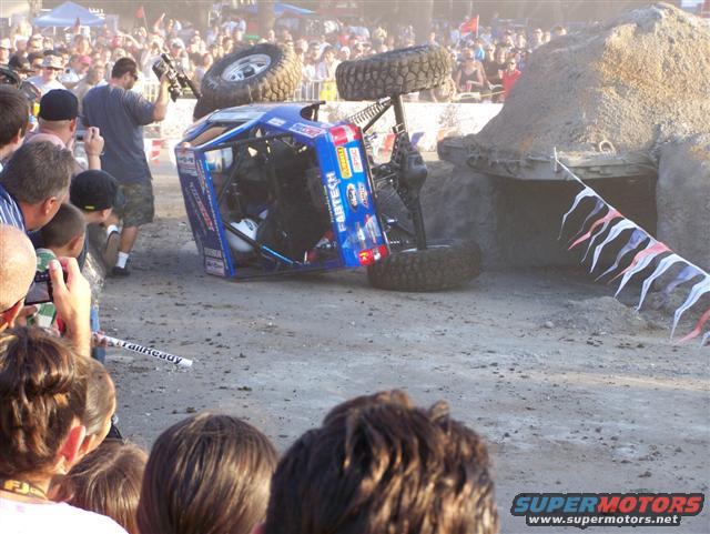 blue off-road truck flipped on its side with large tires, crowd watching
