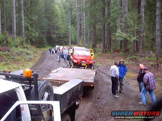 Several off-road vehicles, including a red Jeep, drive down a dirt road in a forest. People stand around a trailer