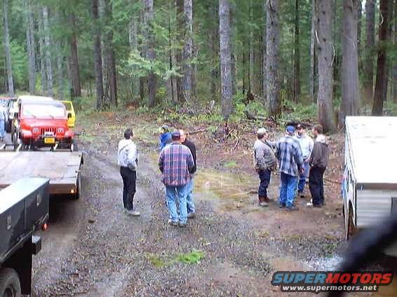 Red Jeep Wrangler on a trailer, front view. A group of people stand around it in a wooded area