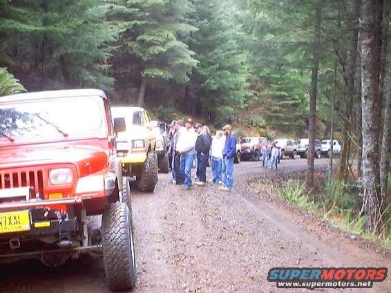 Red Jeep Wrangler front view, yellow Ford Bronco behind it, group of people on dirt road