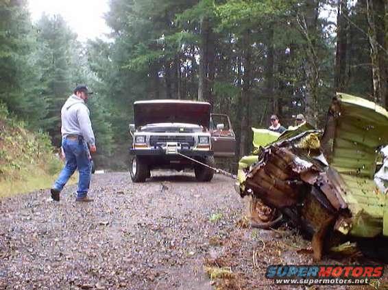 Man walks by a 1980s Ford Bronco being towed by another truck, with a wrecked vehicle in the foreground