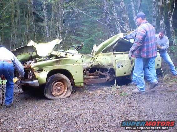 People examining a heavily damaged light green pickup truck with flat tires in a wooded area