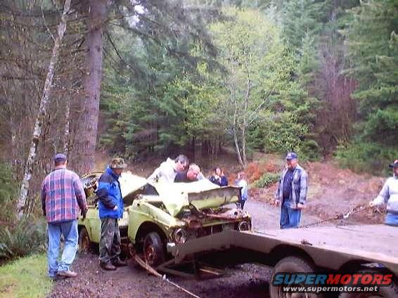 Wrecked light-colored sedan on a trailer, front end heavily damaged, being loaded in a wooded area