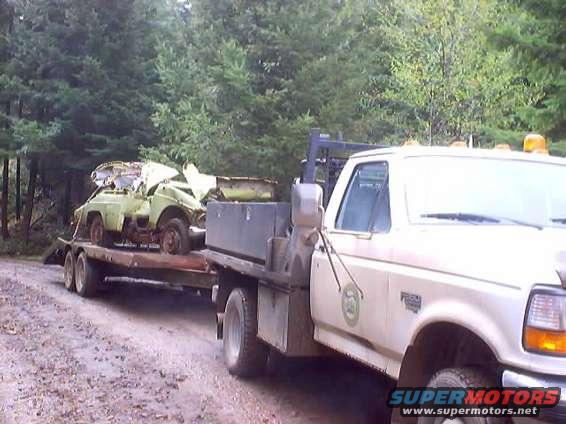 Tow truck hauling a wrecked light-colored car on a trailer. Trees in background
