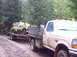 1976 Ford F-100 - A flatbed tow truck transports a heavily damaged vehicle on a trailer through a wooded area.