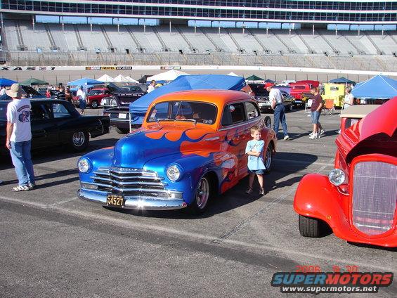 blue and orange hot rod with flames, kid standing next to it, other cars and stadium in background