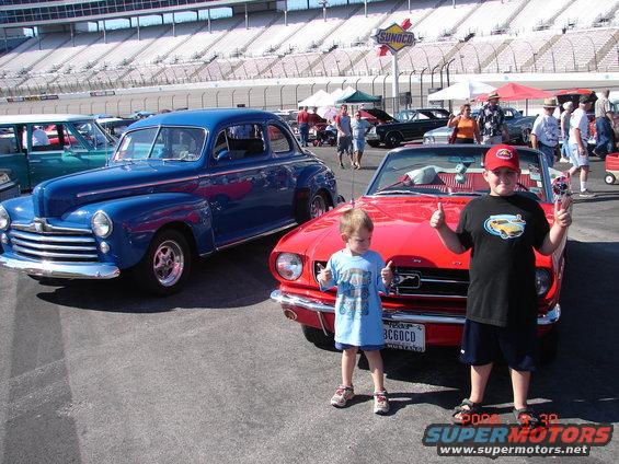 Two boys give thumbs up in front of a blue classic car and a red classic car at a race track with Sunoco signage