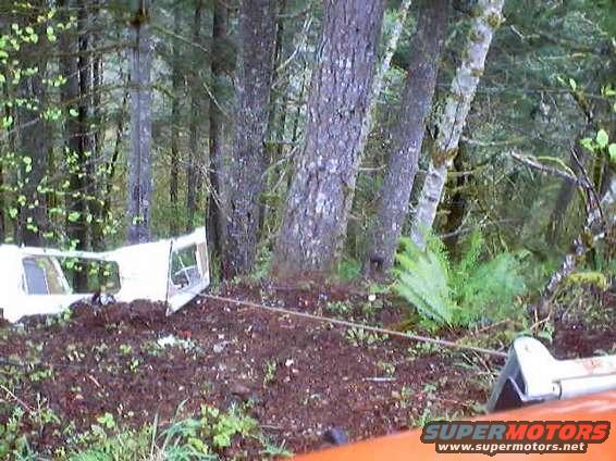 Wrecked white truck cab on its side in a forest, being pulled by a cable. The body style suggests a 1970s pickup