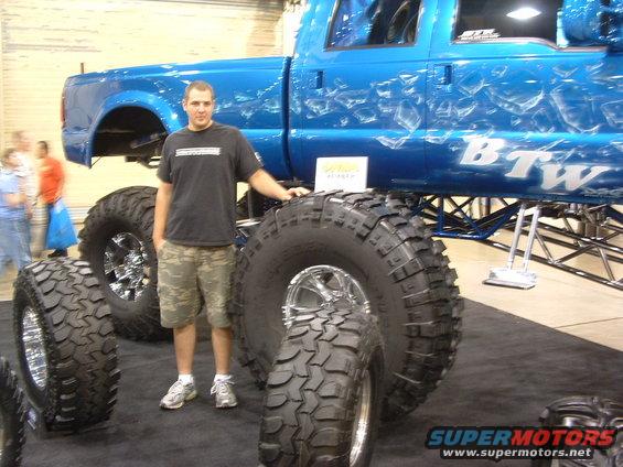 Man next to custom blue pickup truck with large off-road tires and "BTW" graphics