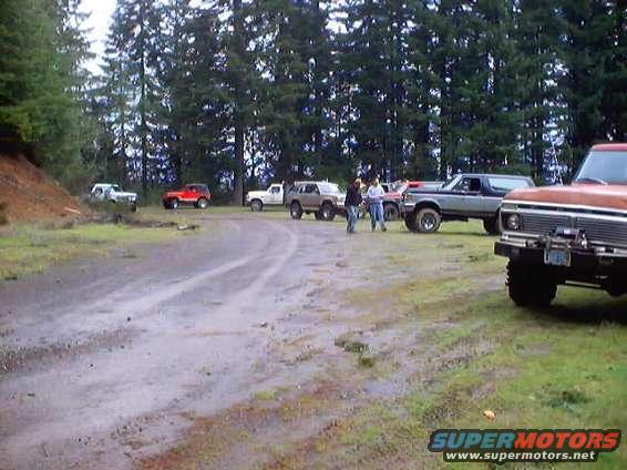 Several trucks and SUVs parked on a muddy dirt road near trees. Some people are standing around