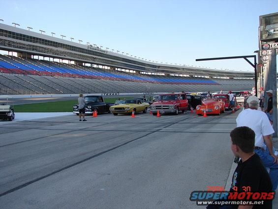 Several classic cars and trucks parked on a racetrack in front of stadium seating