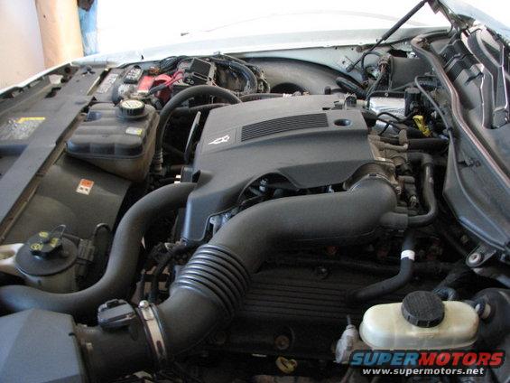 Engine bay with hoses, battery, and engine cover visible. Some text on a fluid reservoir