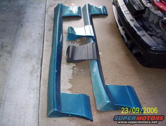 Teal car side skirts and bumper trim pieces on concrete, with a black car bumper visible to the right