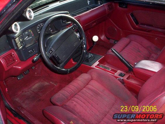 Red interior of a 1990 Ford Mustang with a manual transmission, aftermarket gauges, and worn seats