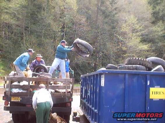 Men are loading old tires into a blue dumpster next to a pickup truck bed full of tires