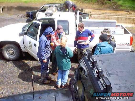 White pickup truck with people gathered around its open driver's door, another vehicle in the background
