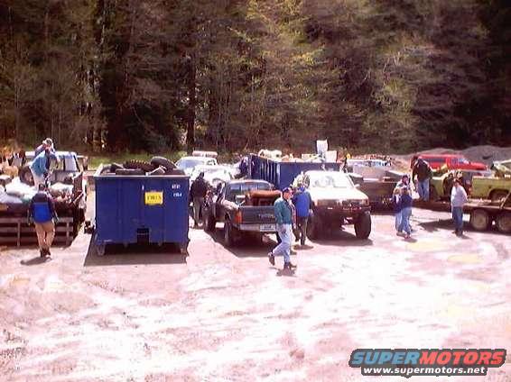 Several trucks and people in a salvage yard with large blue dumpsters