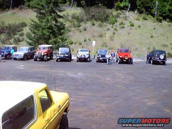 Several SUVs and trucks are parked in a lot with a wooded hill in the background. A yellow vehicle is in the foreground