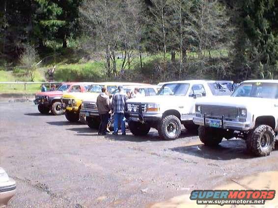 Several pickup trucks parked in a lot with trees in the background. People are standing around the trucks