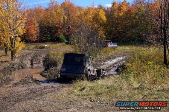 Dark SUV stuck in mud with a pickup truck in the background during autumn