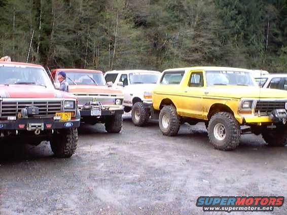 Several lifted Ford Broncos and trucks parked on gravel, with trees in the background