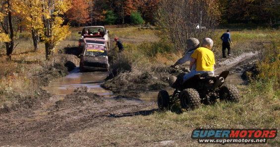 Two people on an ATV watch a truck and another vehicle go through a muddy ditch