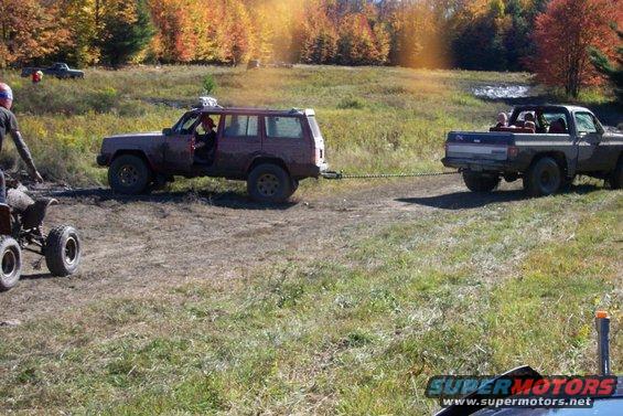 Red SUV being towed by a pickup truck in a muddy field with fall foliage