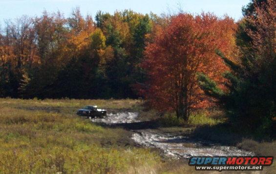 Pickup truck driving through a muddy field with fall foliage in the background