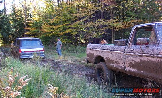Mud-covered 1979 Ford Bronco, rear view, stuck in woods with a tow rope attached to a mud-covered Ford F-Series truck