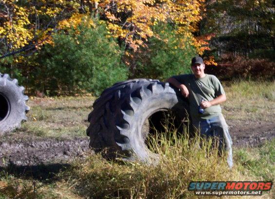 Man in green t-shirt and jeans leans on a large tractor tire in a grassy field with autumn trees in the background. Another large tire is visible to the left