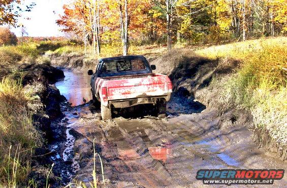 Red pickup truck stuck in muddy ditch with fall foliage in background