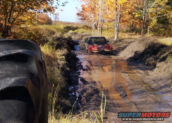 Red pickup truck stuck in a muddy rutted path with trees and fall foliage in background. Large tire in foreground