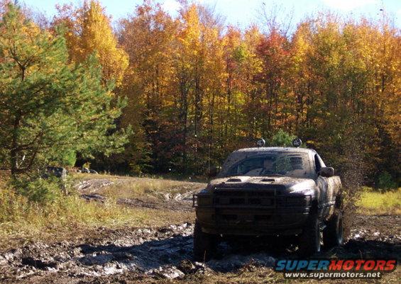 Mud-covered pickup truck driving through a muddy trail with fall foliage in the background